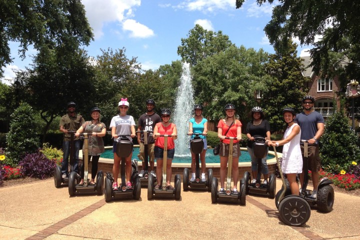 Guests smiling on Segways in front of a beautiful water fountain. Green lush trees are surrounding the water fountain.