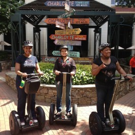 Three women smiling on Segways in front of a directions sign