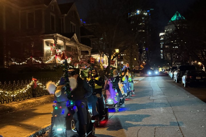 People on decorated scooters in holiday-themed outfits ride down a street at night.