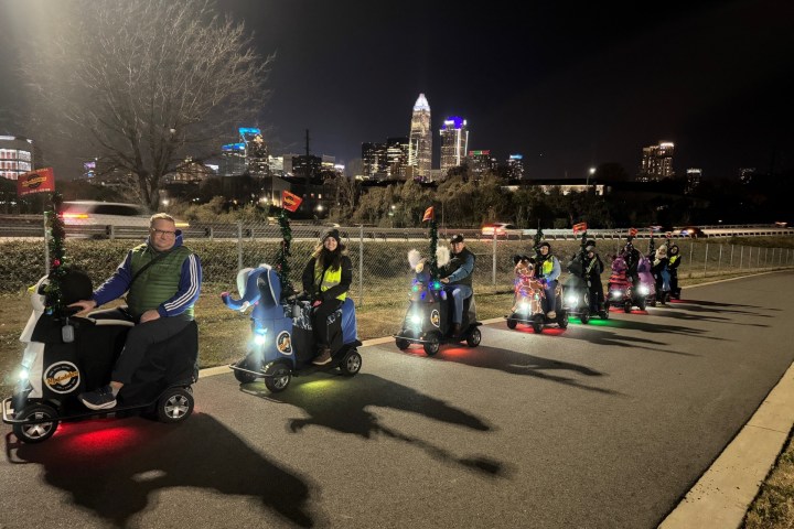 Group of people on scooters decorated with lights, city skyline in background at night.