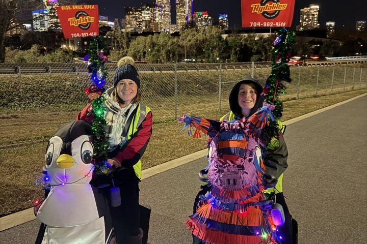 Two people riding scooters decorated as a penguin and llama, with city skyline in the background at night.
