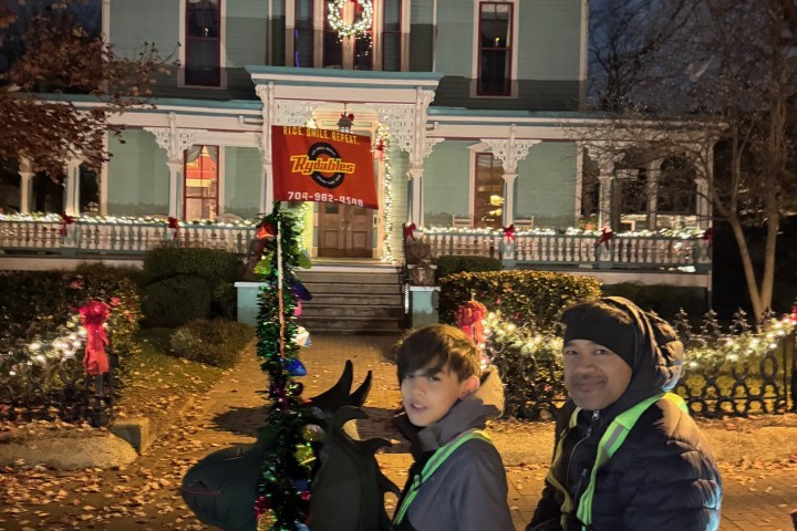 Two people on a decorated ride-on reindeer in front of a festive house at night.